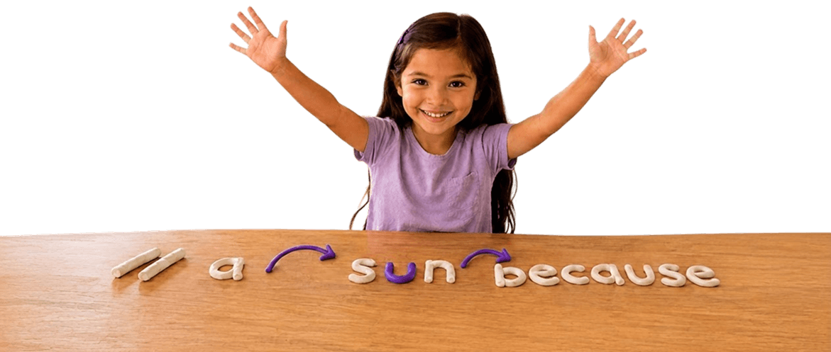 A smiling child raising both hands over a table with clay letters spelling reading practice words