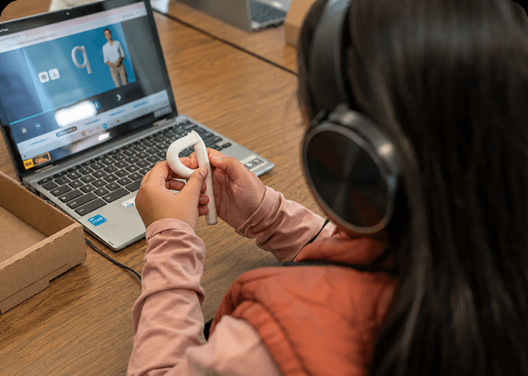 A student wearing headphones shaping a clay letter beside a laptop lesson